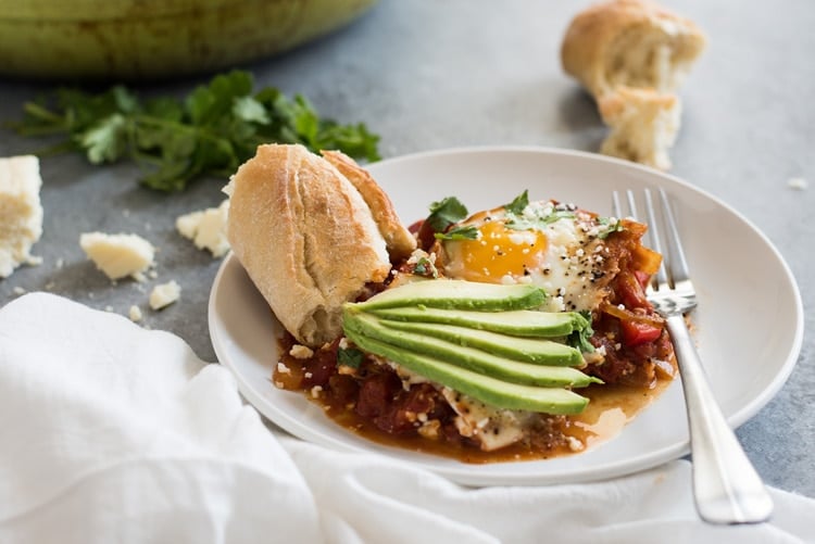 Shakshuka on a white place with avocado slices and fresh crusty bread.