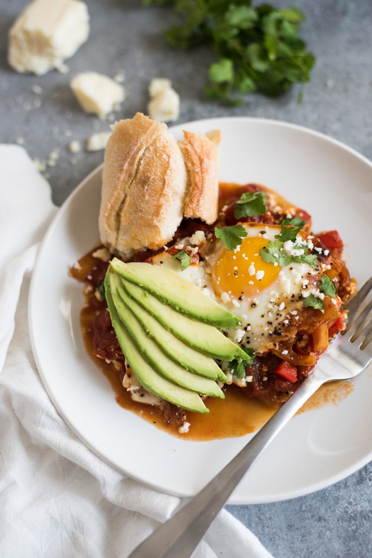 Chipotle Shakshuka recipe on a white plate topped with avocado slices and a side of fresh bread.