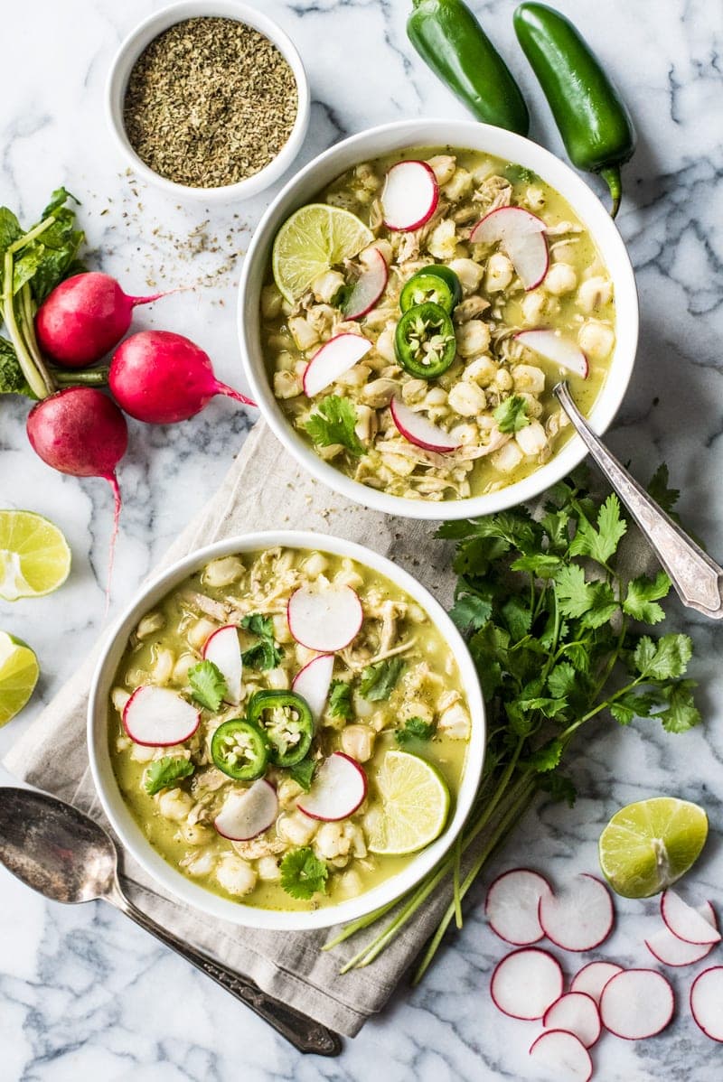 Chicken pozole recipe in two white bowls topped with cilantro, limes and radishes.