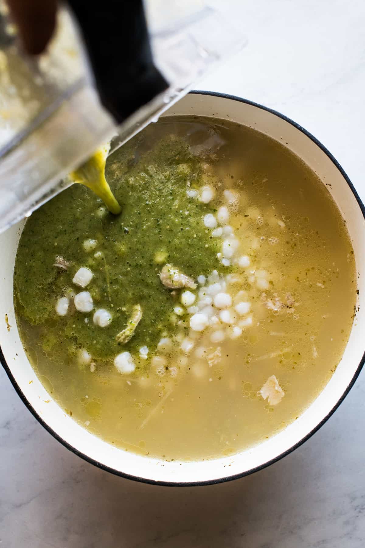 Green chile sauce being poured into a large pot with broth and hominy for pozole verde.