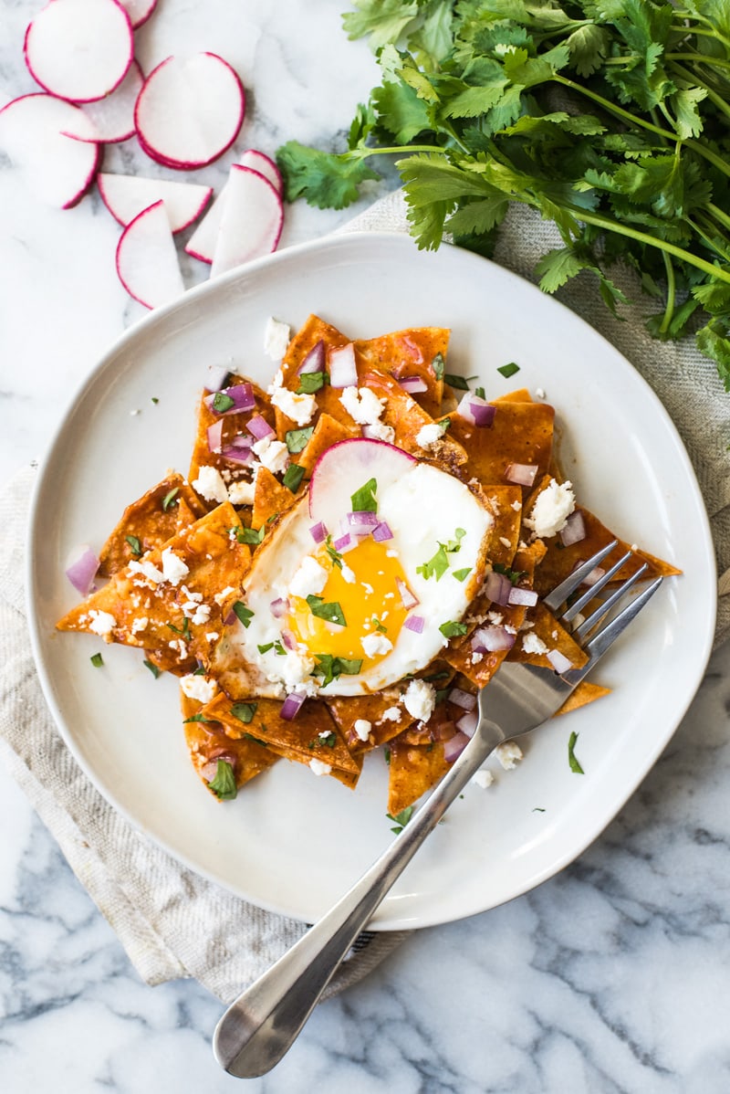 Chilaquiles on a white place topped with a sunny side up egg, chopped cilantro, red onions and cotija cheese.