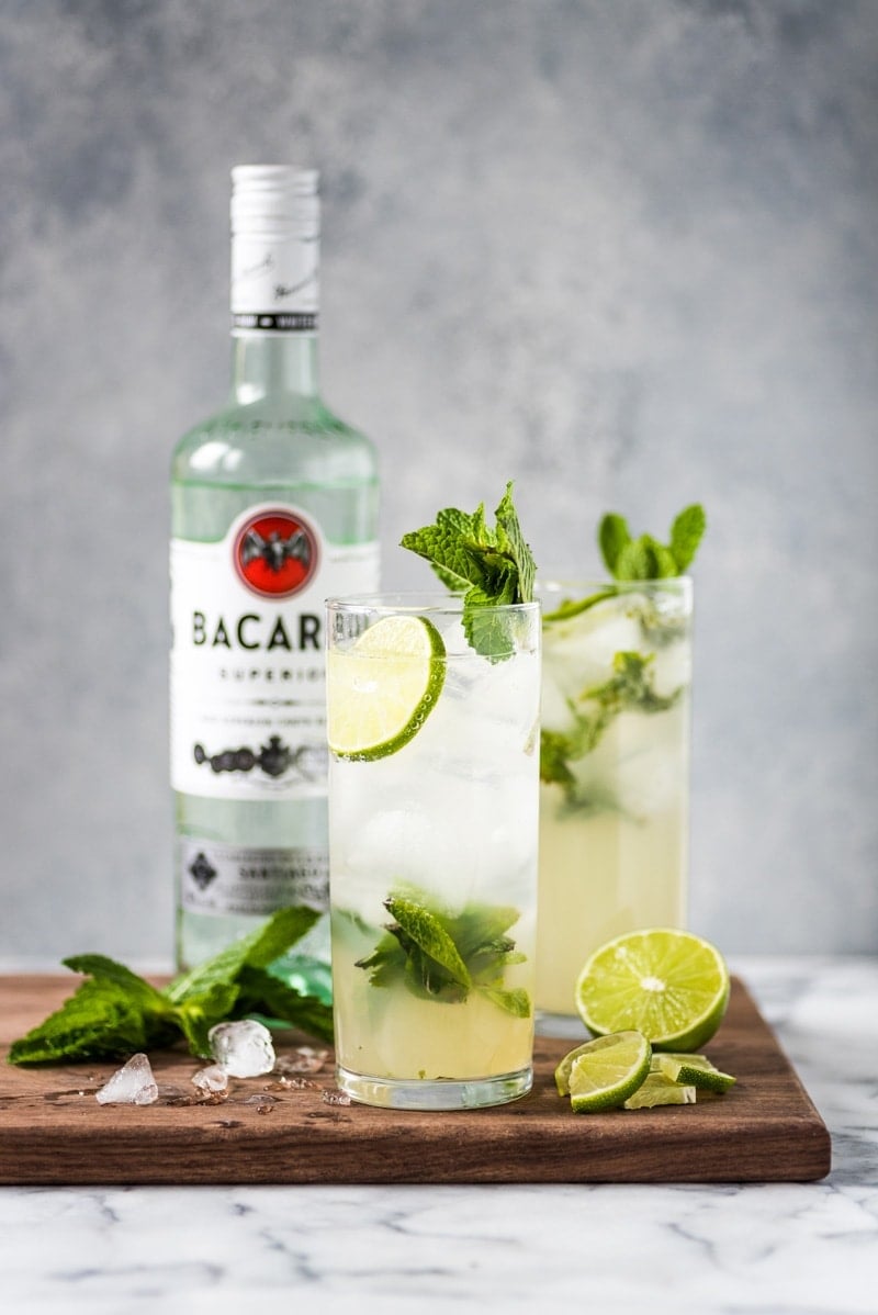 Two glasses of mojitos topped with fresh mint leaves and limes.