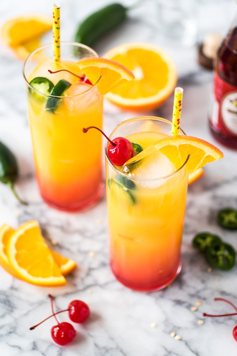 2 glasses of Tequila Sunrise cocktail topped with maraschino cherries and an orange slice.