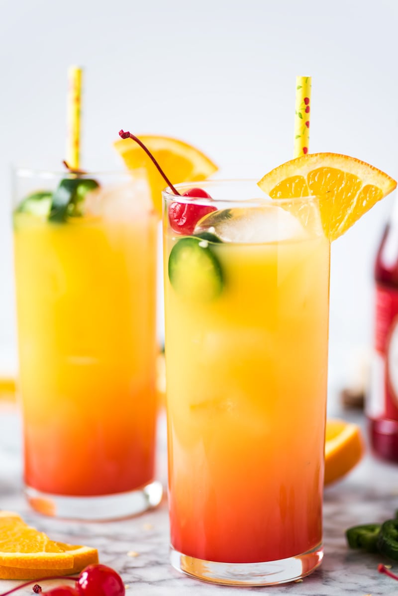 A Tequila Sunrise made with orange juice, tequila and grenadine. Garnished with an orange slice, maraschino cherries and jalapeno slices.