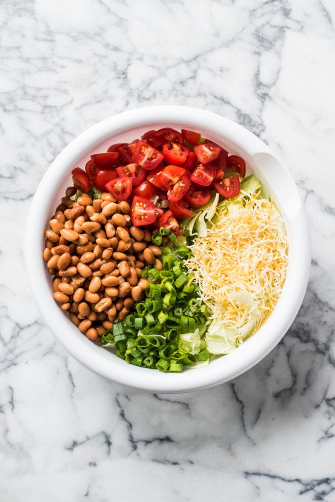 A white mixing bowl filled with chopped tomatoes, pinto beans, iceberg lettuce, cheese and green onions for taco salad.