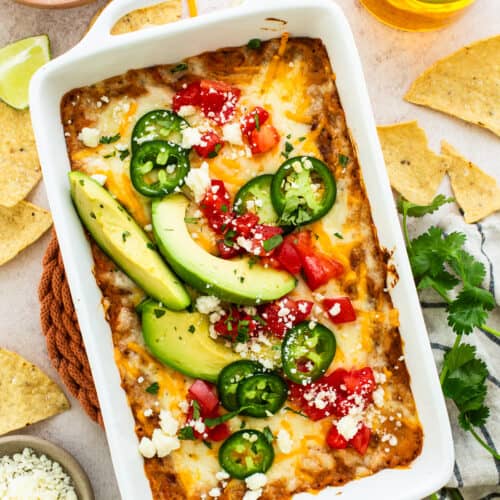 Bean dip topped with melted cheese and garnished with diced tomatoes, cotija cheese, sliced peppers, and avocados.