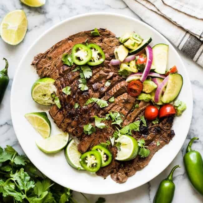 Thinly sliced carne asada on a white plate served with fresh lime wedges and jalapenos.