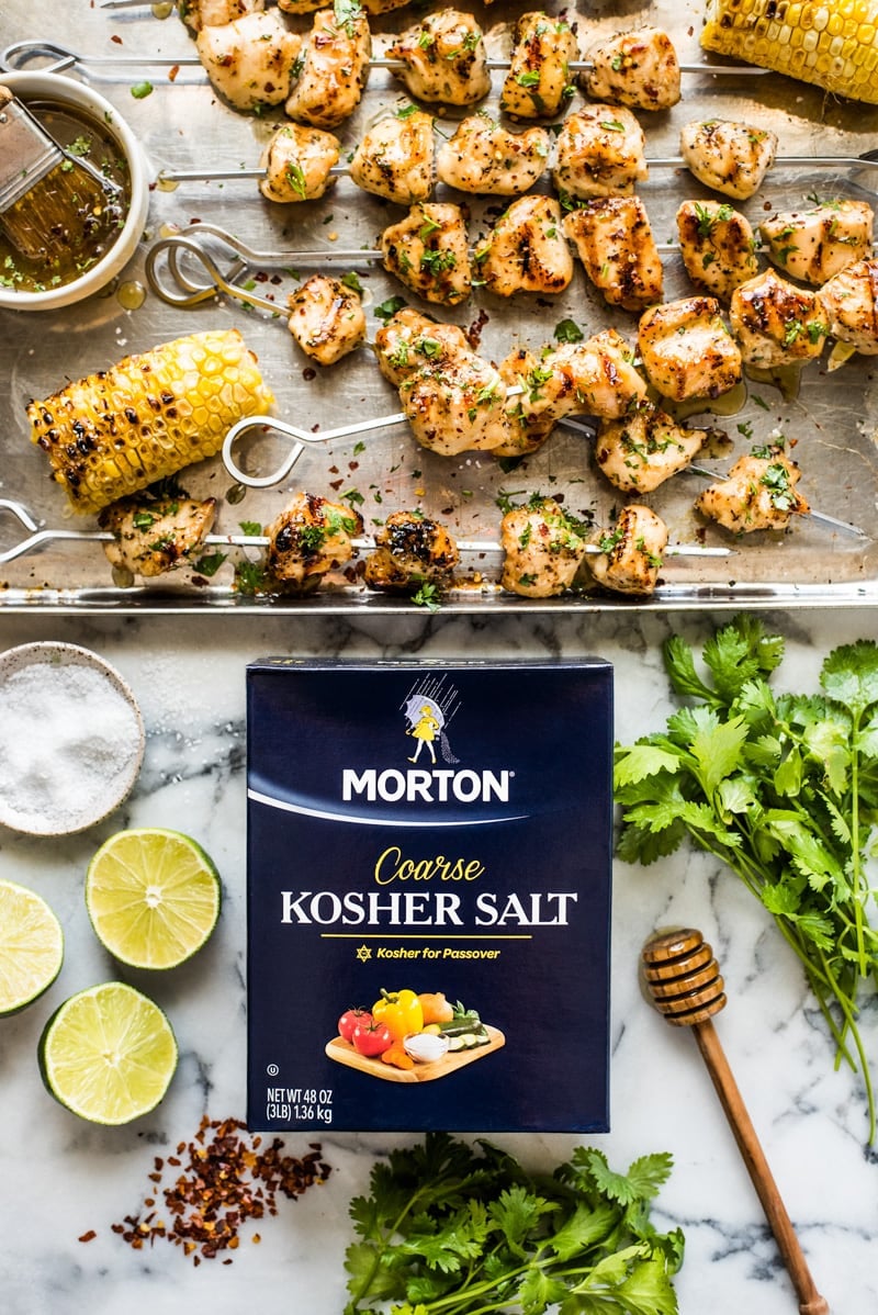 Chicken kabobs on a baking sheet next to Morton Coarse Kosher Salt, cilantro, limes and red pepper flakes.