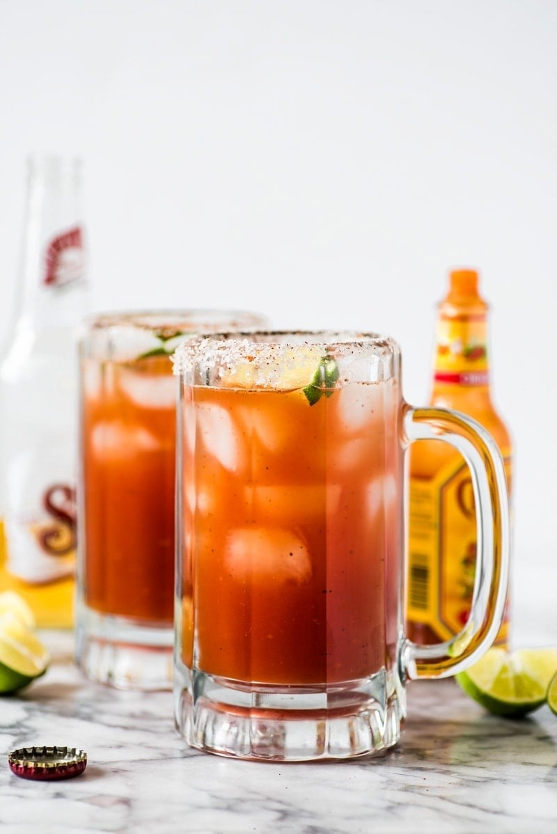 A Michelada cocktail on a marble countertop next to lime wedges, Mexican beer and hot sauce.