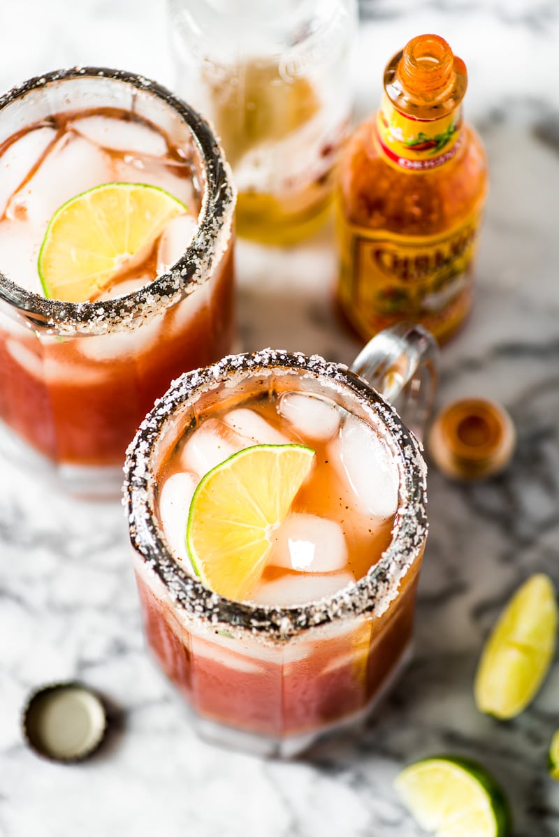 Two micheladas in beer pint glasses topped with lime wedges next to hot sauce.