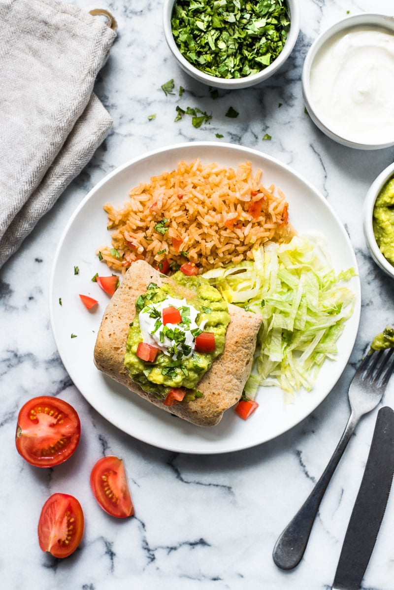 Baked chicken chimichanga topped with guacamole and sour cream on a plate with shredded lettuce and mexican rice.