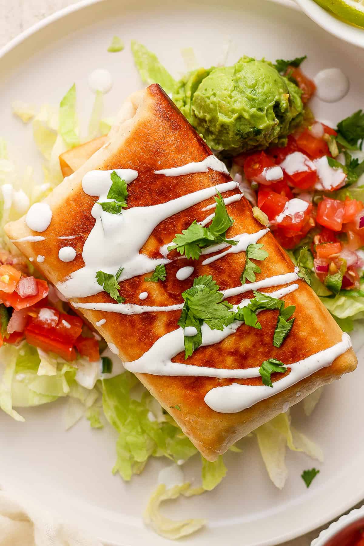 Chimichangas filled with shredded chicken, beans, and cheese, and garnished with a drizzle of Mexican crema or sour cream, cilantro, and served with pico de gallo and guacamole.
