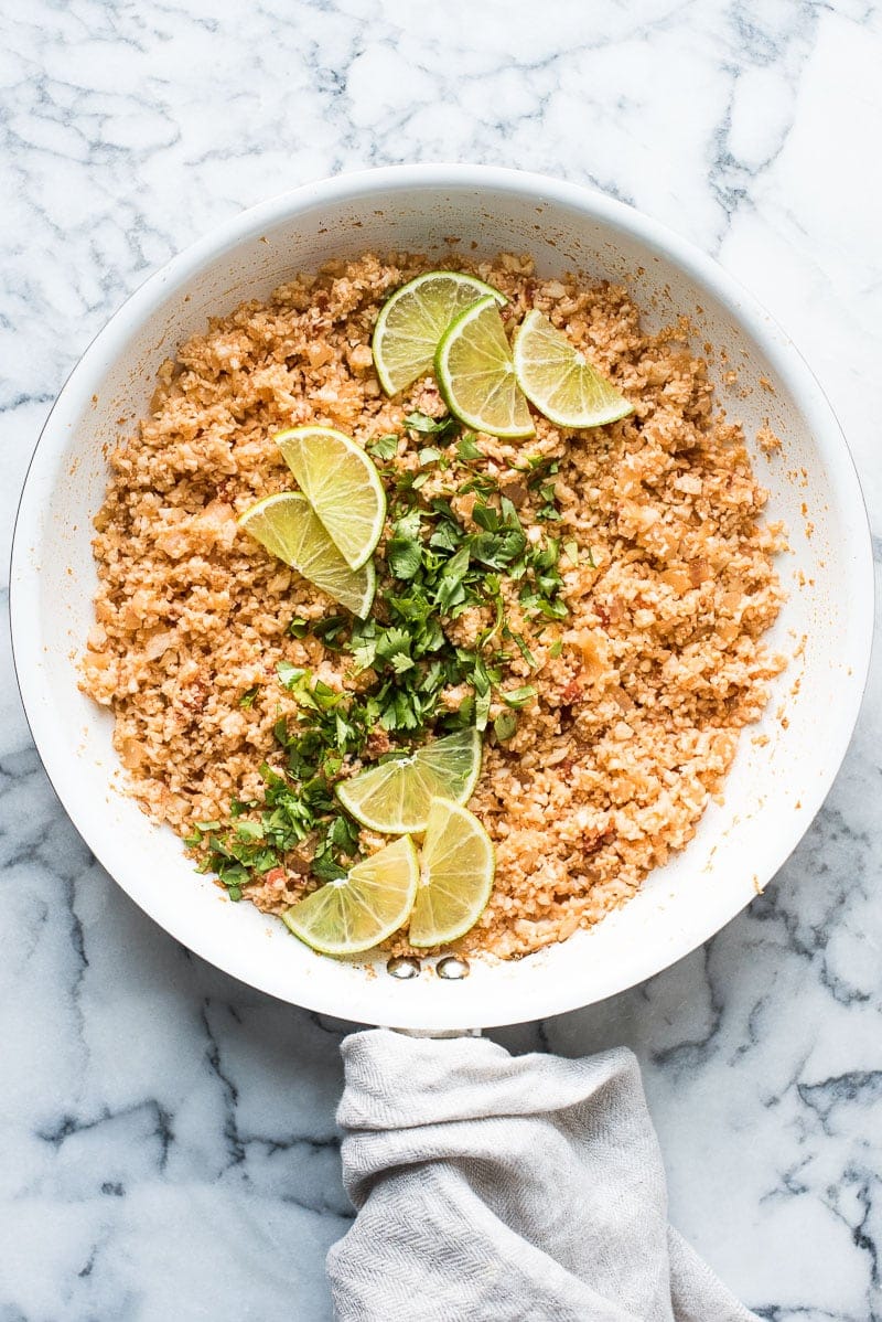 Mexican cauliflower rice in a large skillet garnished with cilantro and lime wedges.