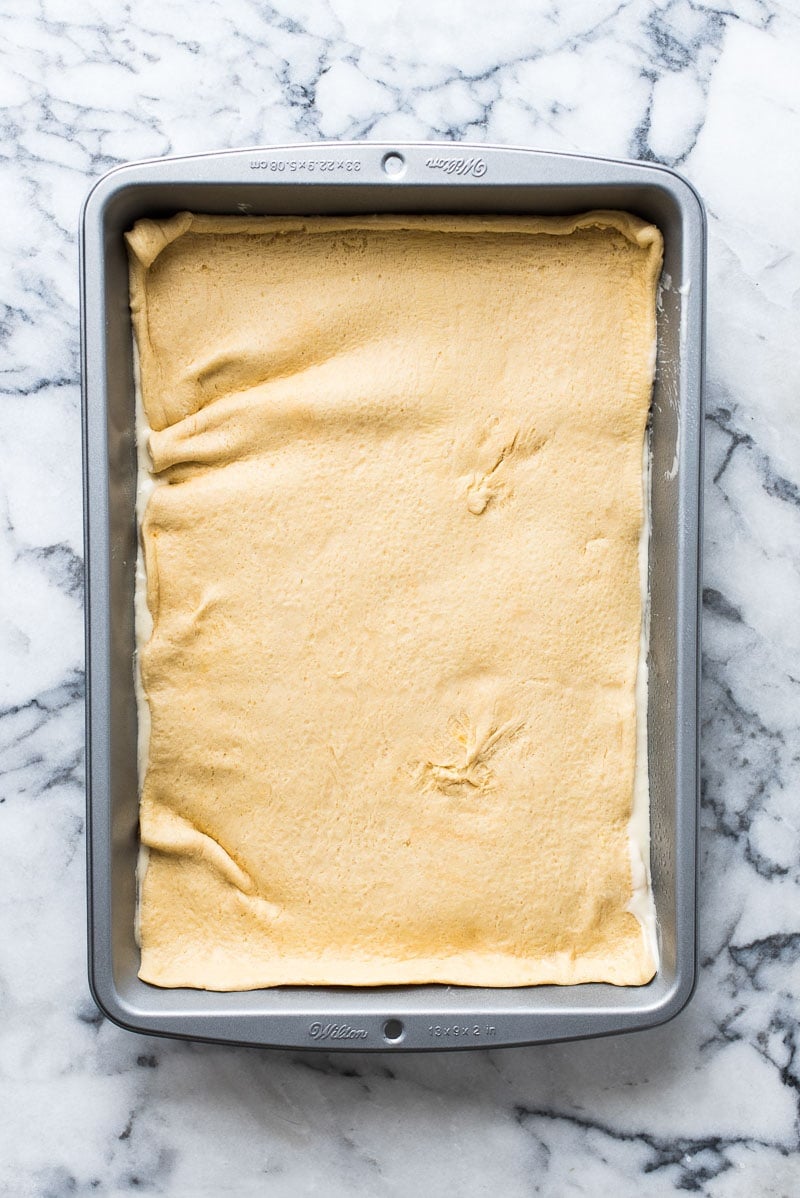 Step three in making sopapilla cheesecake bars. Rectangular 9x13 inch baking dish with crescent roll dough layered on top of cheesecake filling.