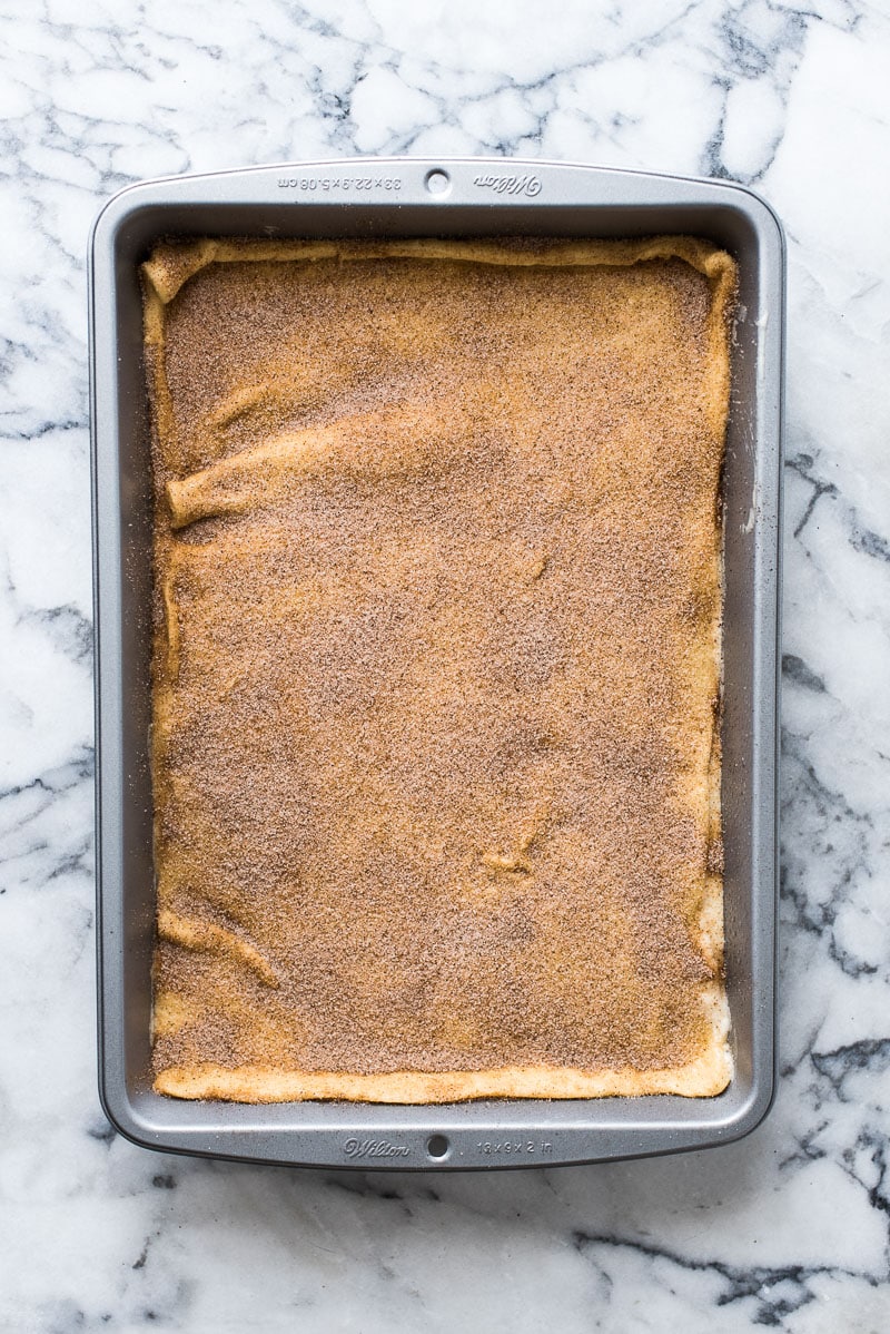 Step four in making sopapilla cheesecake bars. Rectangular 9x13 inch baking dish with crescent roll dough topped with cinnamon sugar.