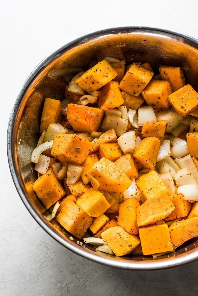 Cubed and peeled butternut squash, chopped onions, and fresh herbs and spices in a large mixing bowl
