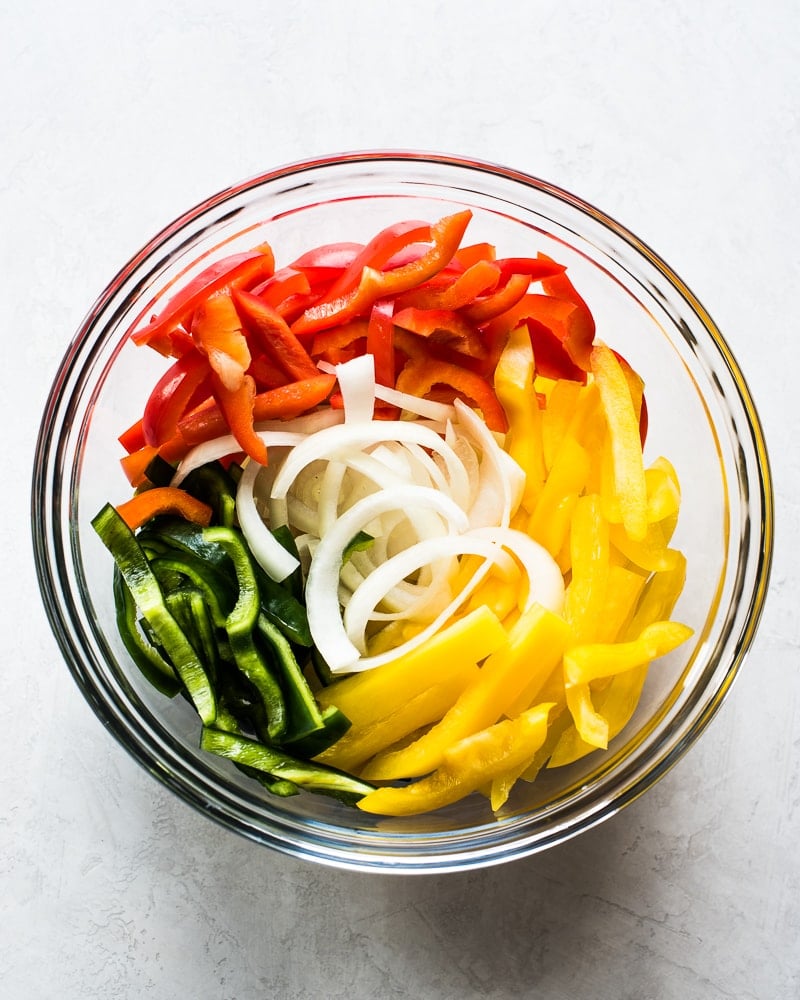 Sliced red, green and yellow bell peppers and onions in a clear bowl.