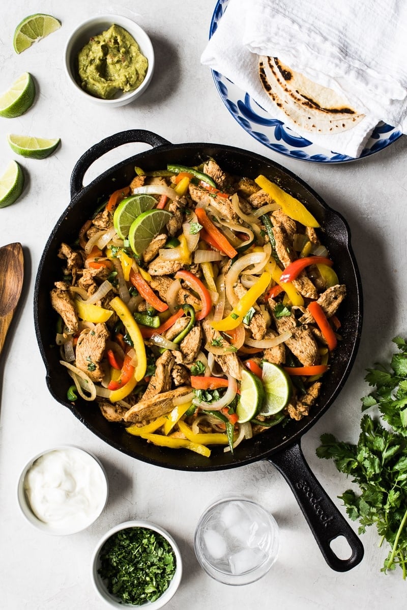 Chicken fajitas with peppers and onions in a black cast iron pan with Mexican ingredients around it.