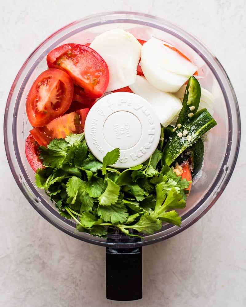 Raw salsa ingredients in a food processor - onions, tomatoes, cilantro, garlic, jalapenos.