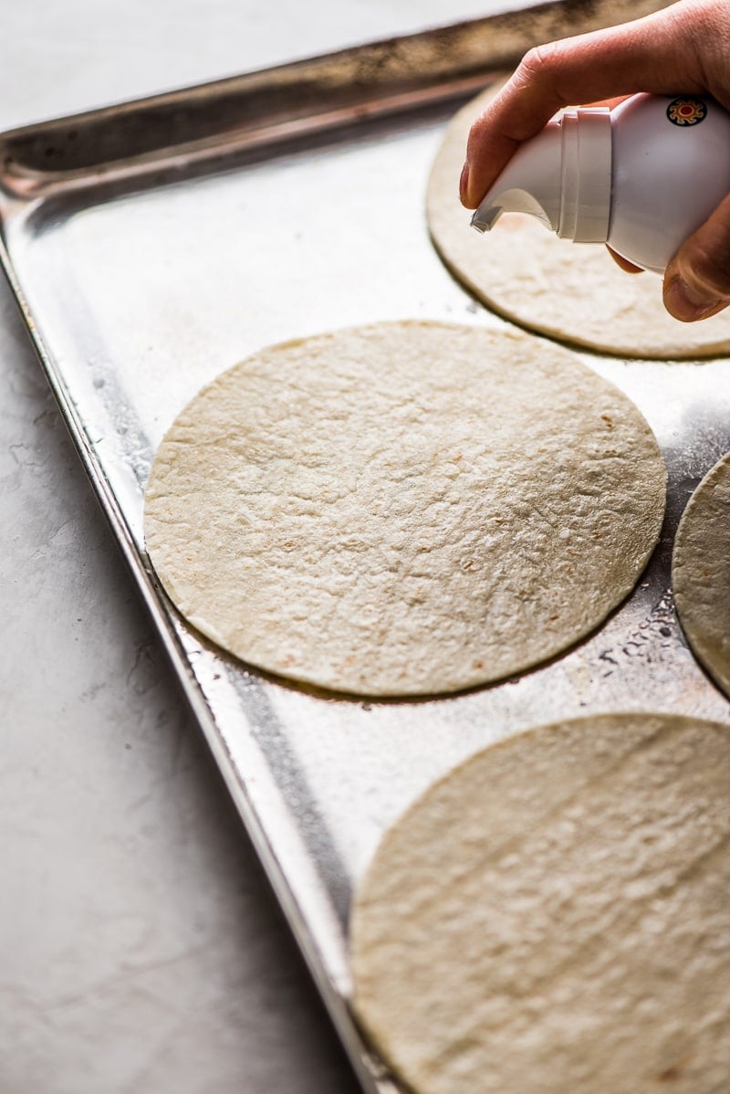 Corn tortillas being sprayed with avocado oil on a large baking sheet.