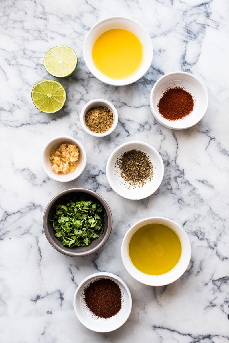 Steak fajita marinade ingredients on a table - olive oil, lime juice, orange juice, garlic, oregano, cumin, smoked paprika and cilantro.