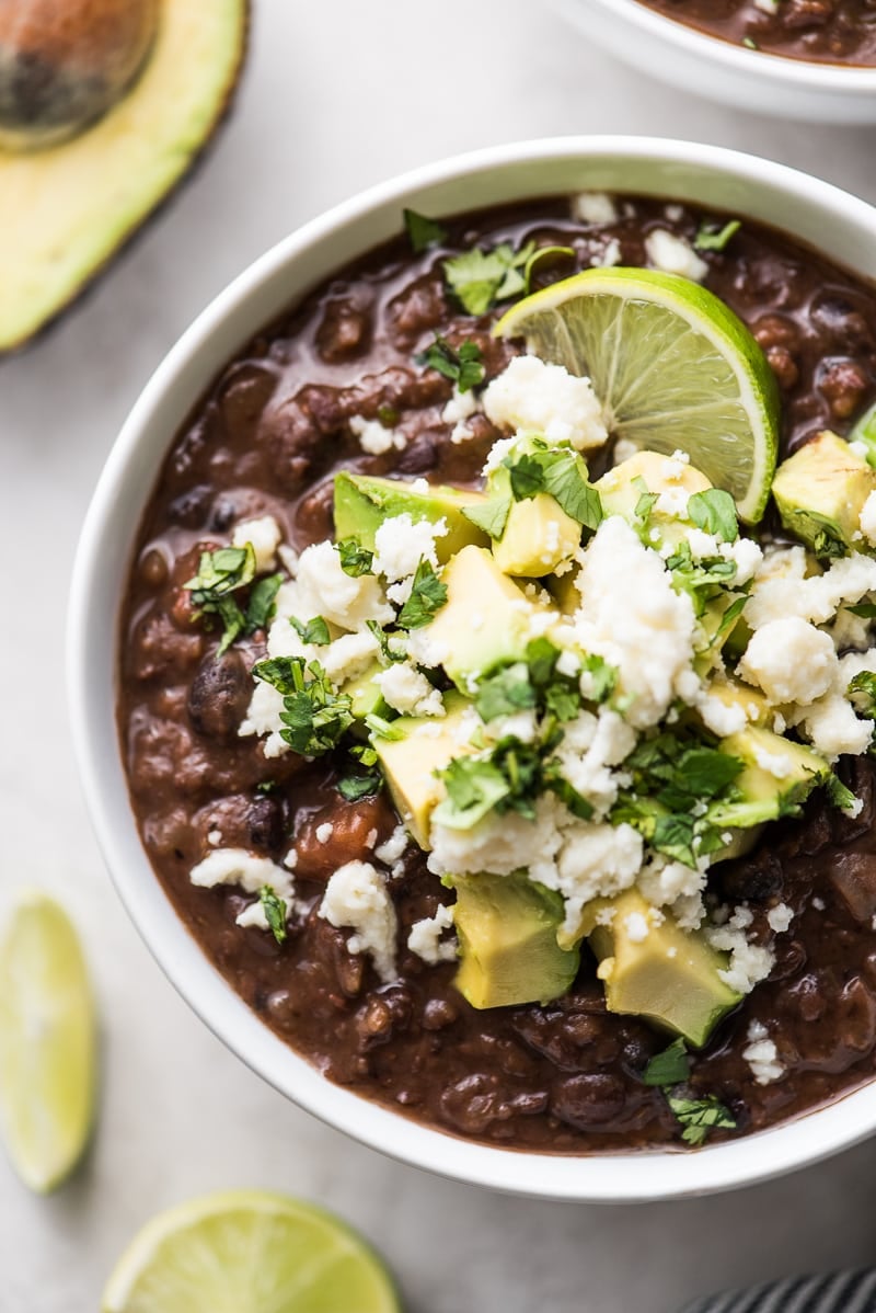 Vegan Black Bean Soup in a bowl topped with avocados, cheese, cilantro and a lime wedge.