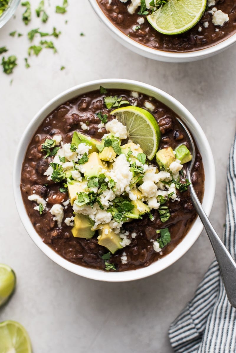 Black Bean Soup recipe in a bowl topped with avocados, cheese, cilantro and a lime wedge.
