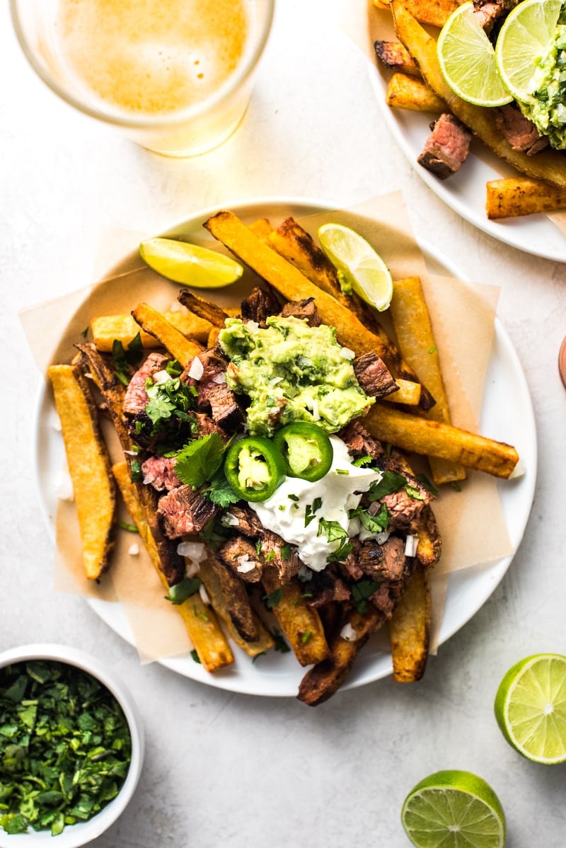Carne asada fries on a white table next to a glass of beer and limes.