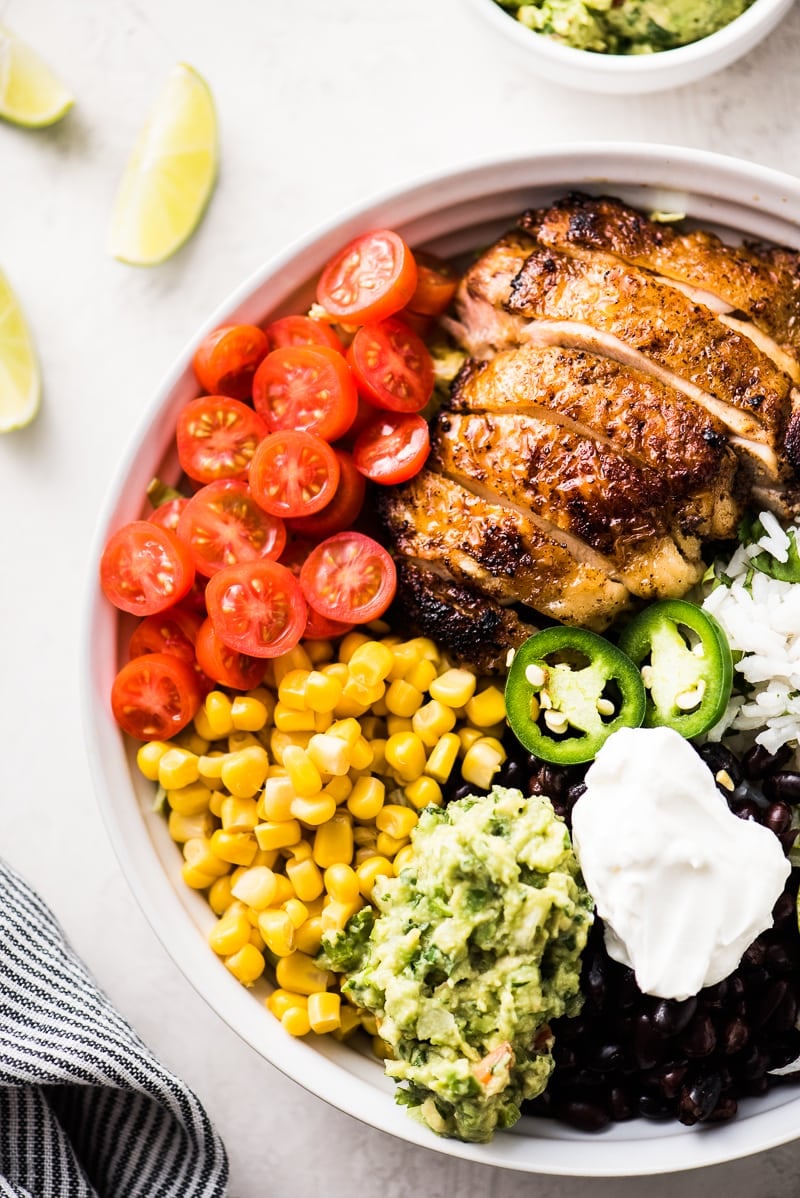 A white bowl filled with chicken, cilantro lime rice, black beans, tomatoes, guacamole and sour cream.