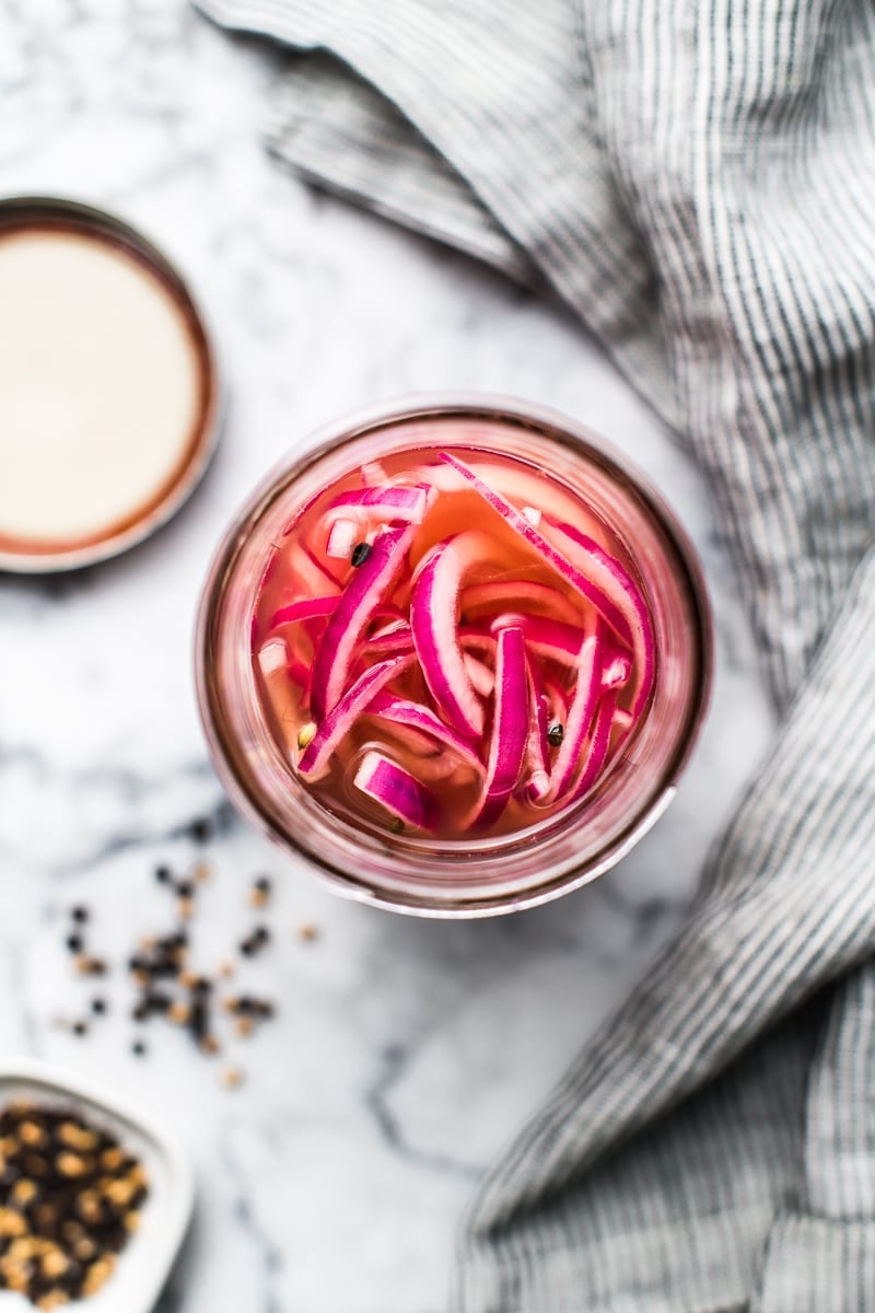 Pickled red onions in a glass jar on a marble table with a kitchen towel and whole black peppercorns strewn on the side.