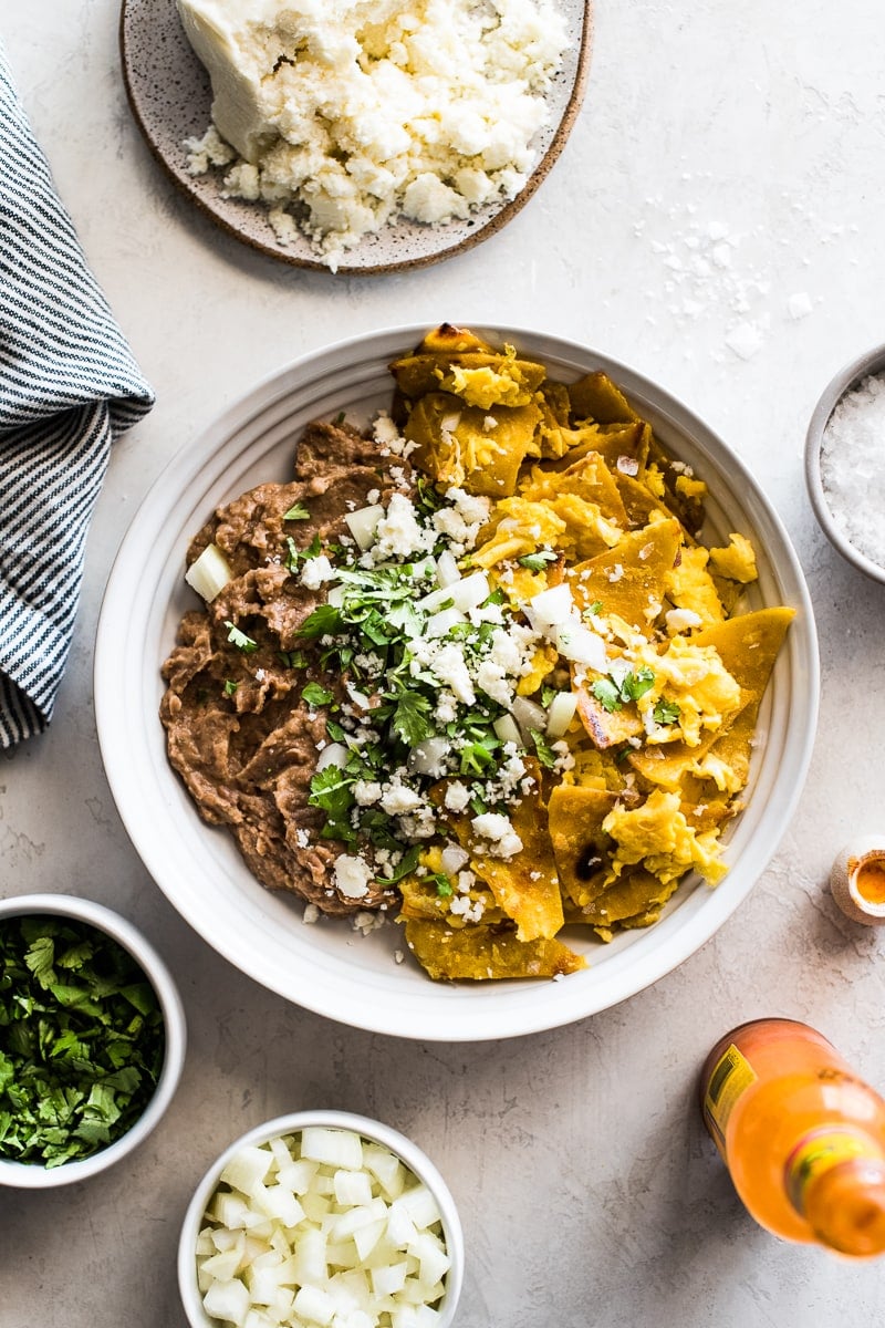 A white bowl filled with migas and refried beans topped with cilantro, onions and queso fresco.