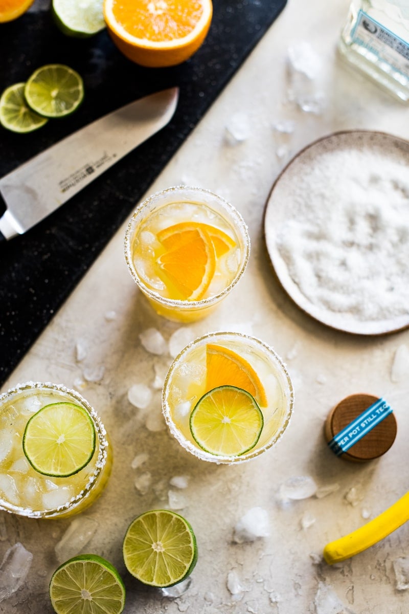 Skinny Margaritas on a table with salt, limes, tequila, oranges and ice cubes.