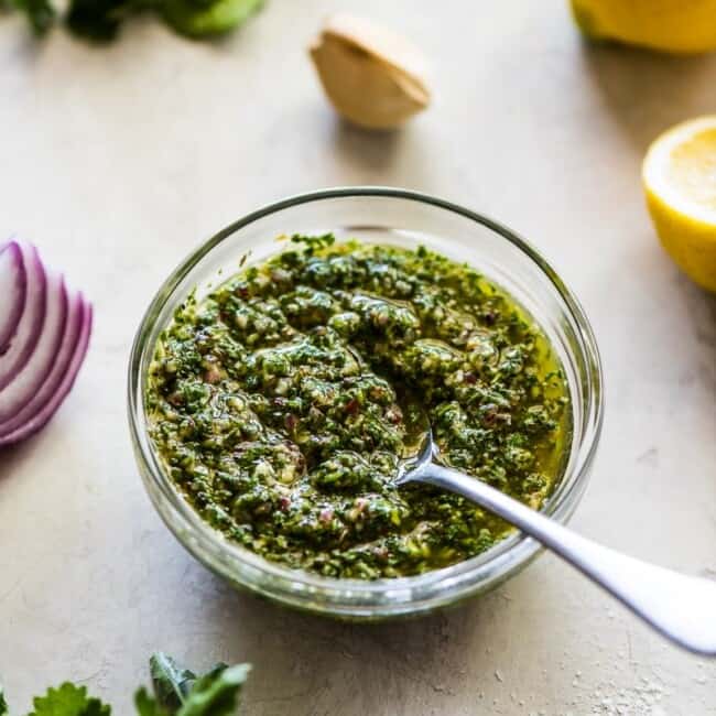 Chimichurri in a small glass container alongside fresh cilantro, parsley and red onions.