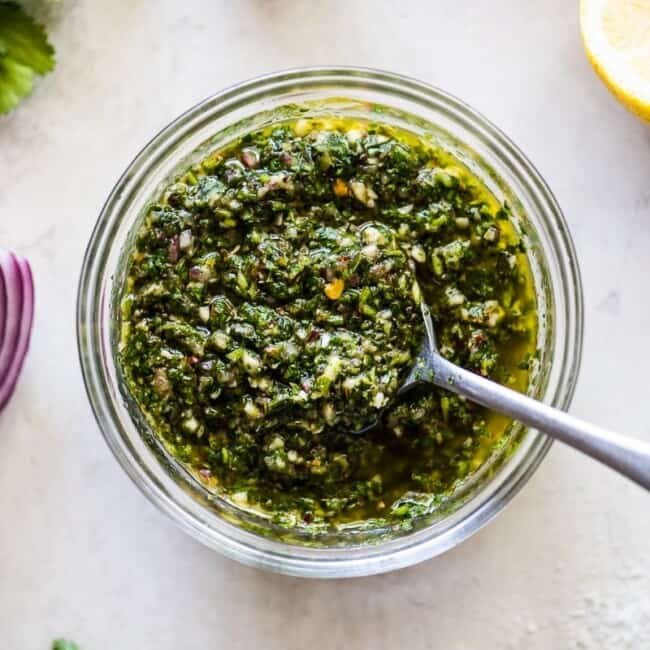 Chimichurri sauce in a clear bowl surrounded by parsley, red onions, cilantro, garlic and lemon juice.