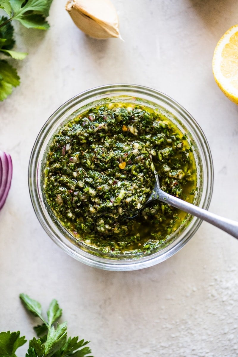 Chimichurri sauce in a clear bowl surrounded by parsley, red onions, cilantro, garlic and lemon juice.