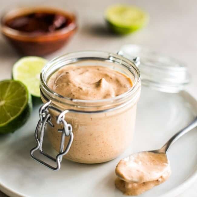 Chipotle sauce recipe in a small glass container surrounded by limes and chipotle peppers.