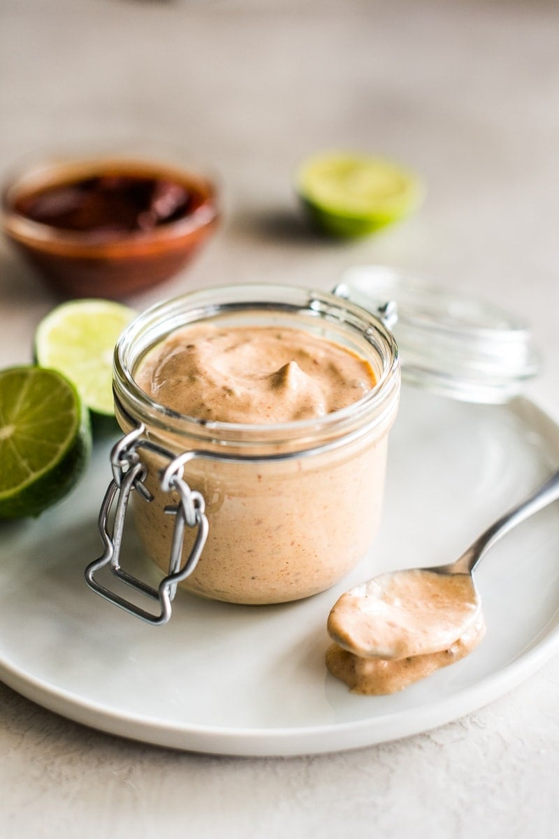Chipotle sauce recipe in a small glass container surrounded by limes and chipotle peppers.