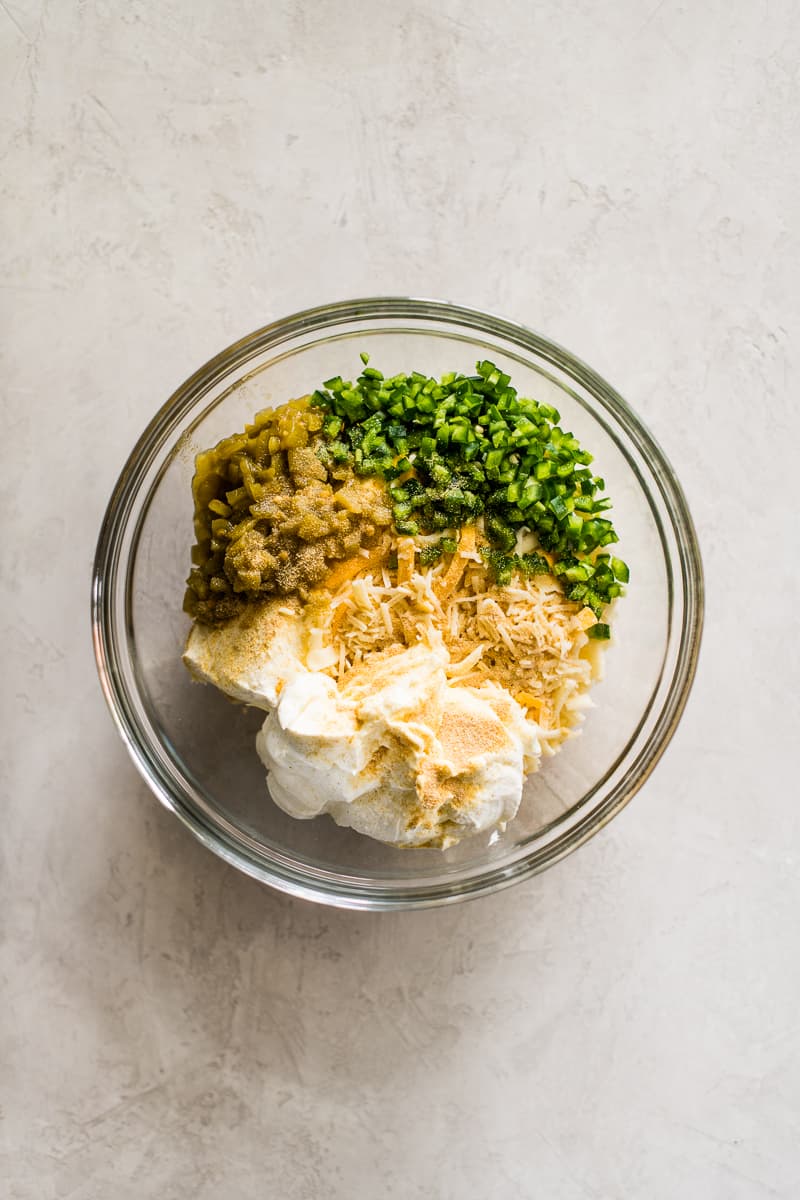 A mixing bowl filled with cream cheese, sour cream, shredded cheese, diced green chiles, jalapenos, and seasonings.