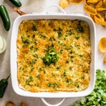 Jalapeno Popper Dip in a white square baking dish surrounded by corn chips, jalapenos, cilantro and margaritas.