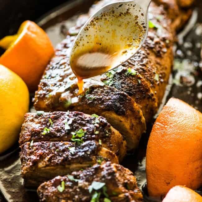 Un cucchiaio che versa il succo di agrumi sul filetto di maiale arrosto in una padella di ghisa nera.