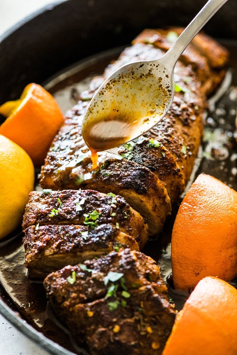 A spoon pouring pork tenderloin oven juices from a black cast iron skillet.