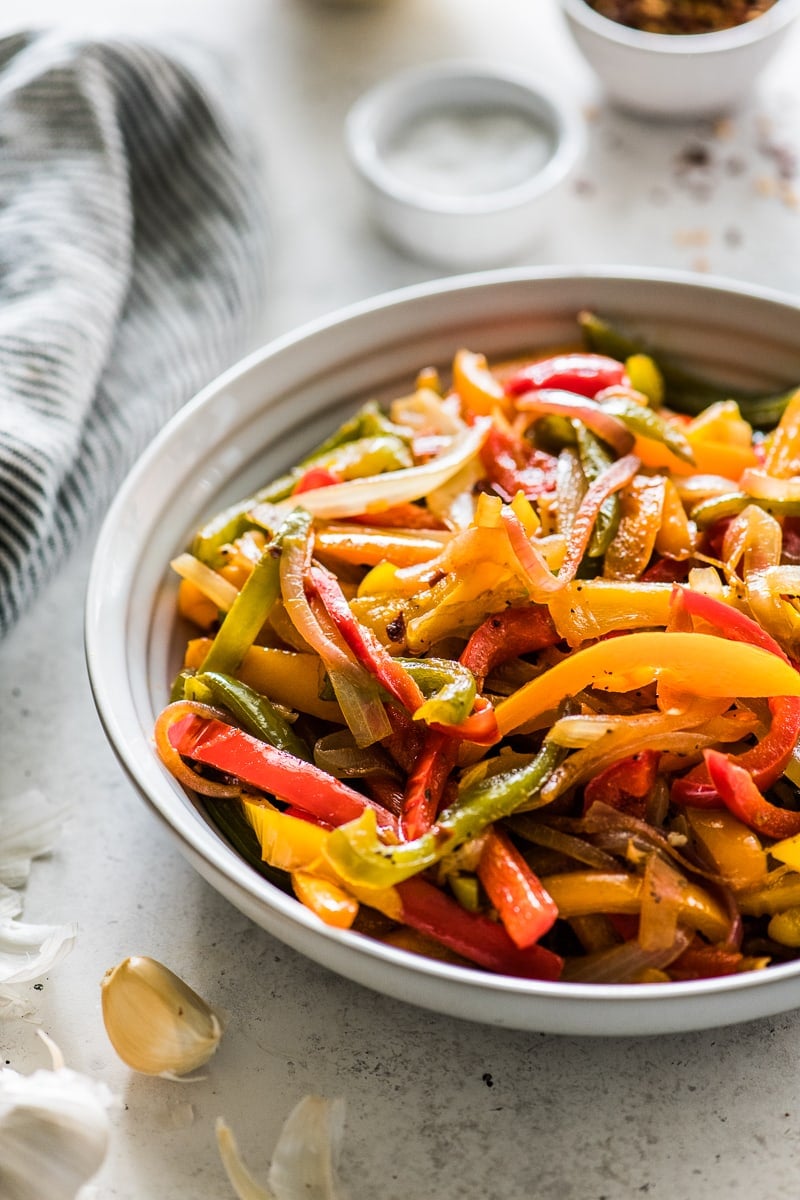 Sauteed peppers and onions in a white bowl next to a clove of garlic and a striped blue and white cloth napkin.