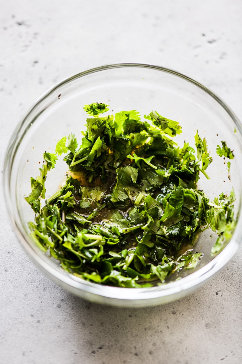 A small clear bowl with chopped cilantro, olive oil, honey, lime juice and spices.