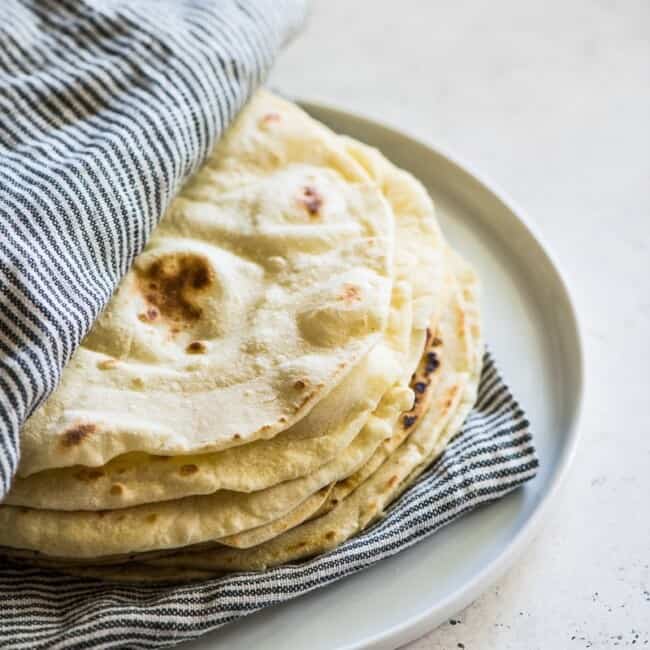 Flour tortillas in between a striped kitchen towel on a white plate.