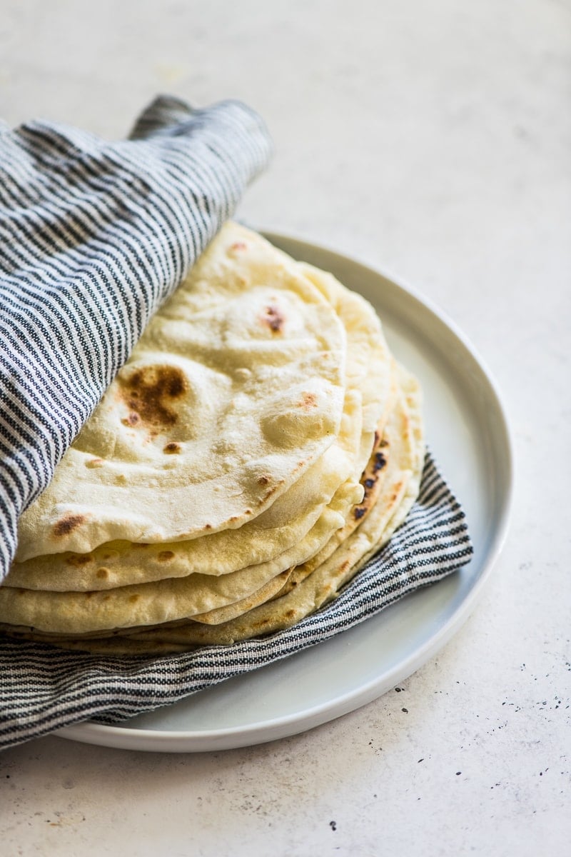 A stack of flour tortillas in between a striped kitchen towel on a white plate.