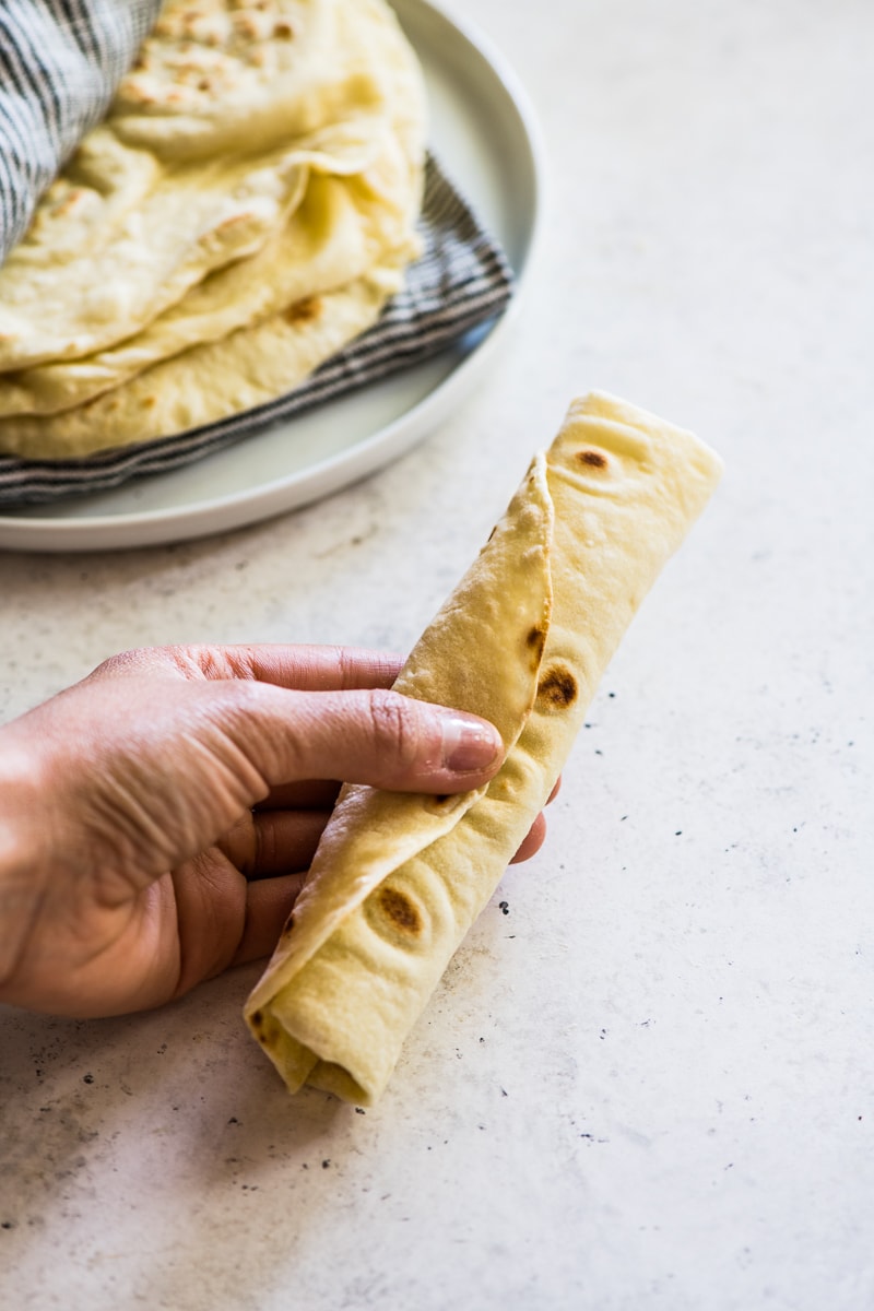 A hand holding a rolled flour tortilla with a stack of tortillas in the background.