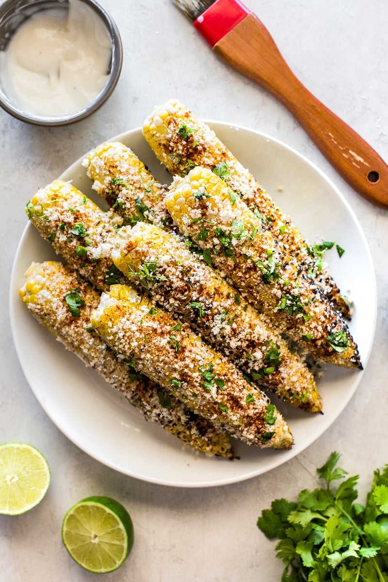 Mexican street corn on a white platter surrounded by limes, cilantro and mayonnaise.