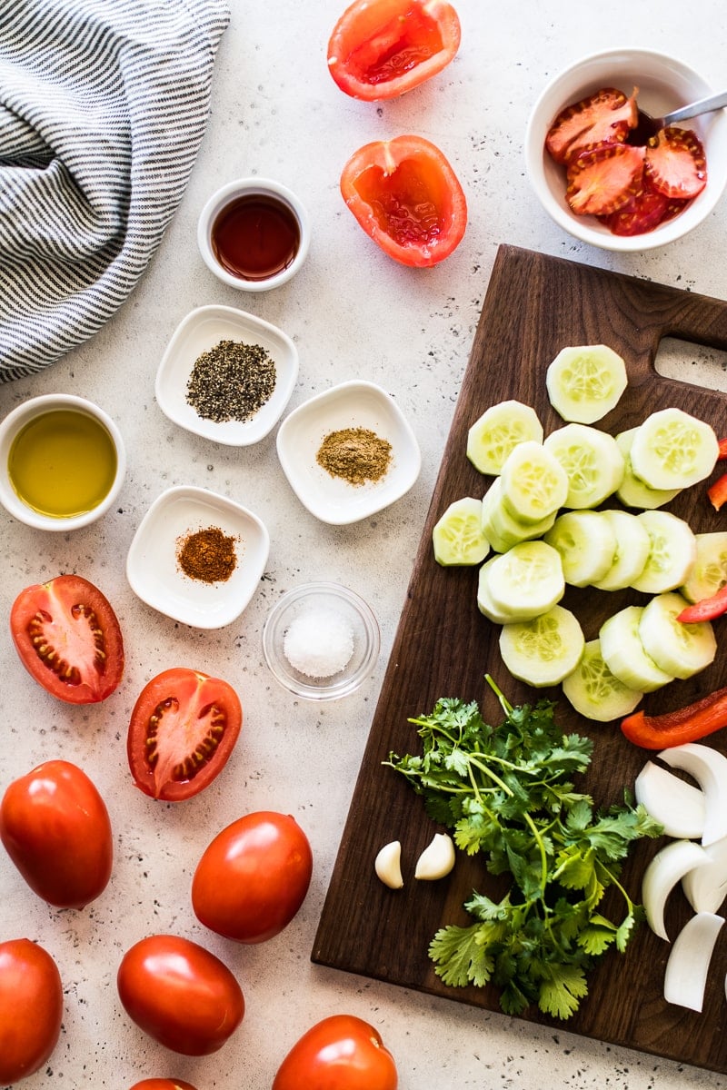 Ingredients for gazpacho on a white and blue table. Includes tomatoes, cucumbers, spices, olive oil, garlic, onions, red bell peppers and red wine vinegar.