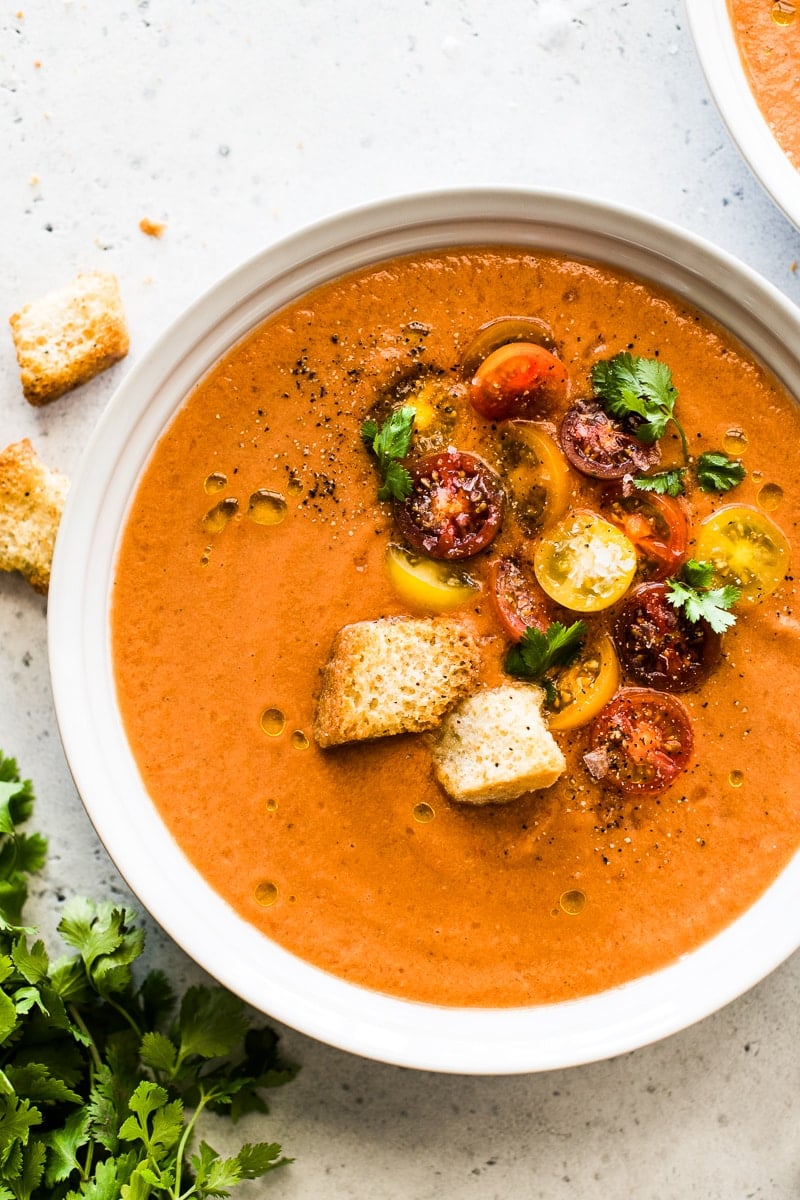 Gazpacho in a white bowl topped with a drizzle of olive oil, cherry tomatoes and croutons.