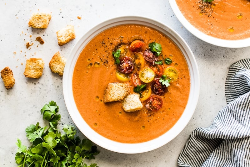 Gazpacho in a white bowl topped with halved cherry tomatoes, croutons, cilantro and olive oil.